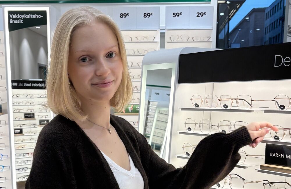 Kuvassa Specsaversin Tiia myymälässä silmälasihyllyn luona.