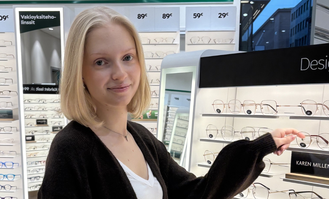 Kuvassa Specsaversin Tiia myymälässä silmälasihyllyn luona.