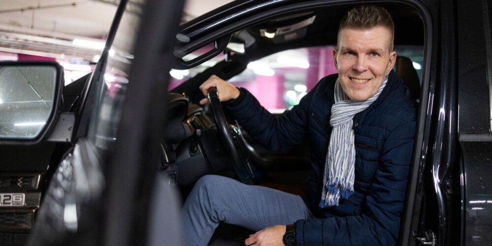Mies istuu autossa ja hymyilee sen ovenraosta.