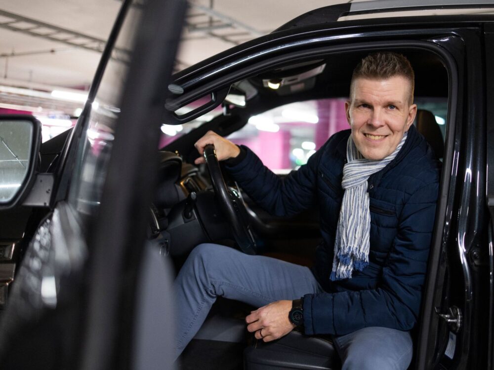 Mies istuu autossa ja hymyilee sen ovenraosta.