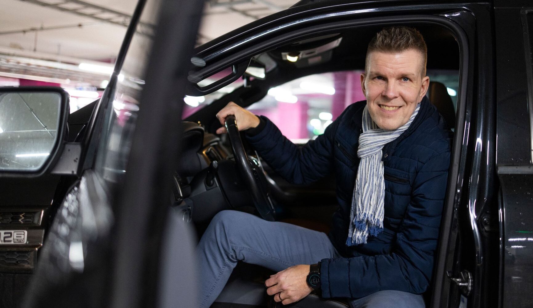 Mies istuu autossa ja hymyilee sen ovenraosta.